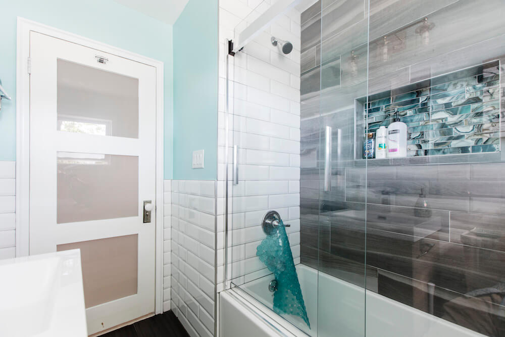 Gradient gray tiled bathwall for bathtub area with glass door and storage niche after renovation
